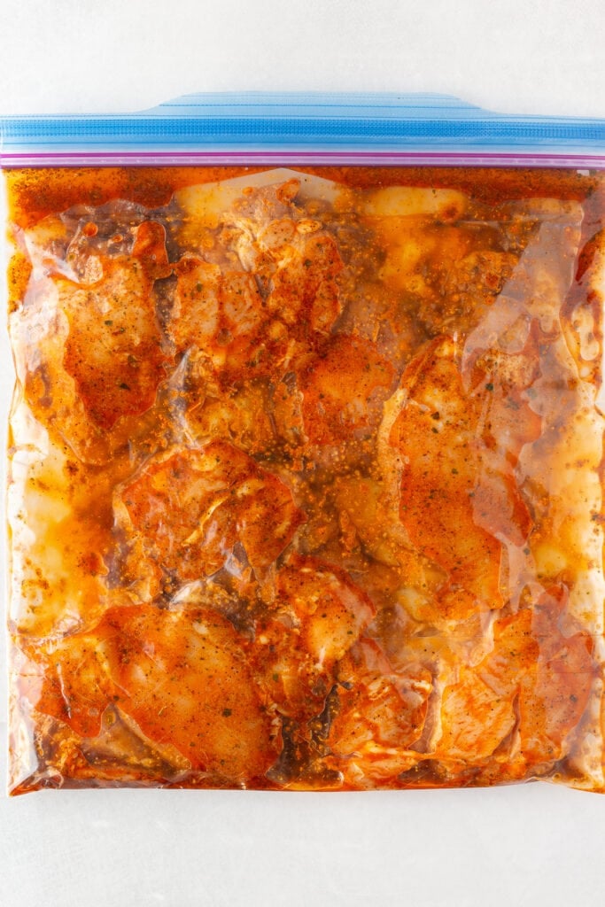 Top down shot of boneless skinless chicken thighs covered in a red marinade in a gallon sized plastic ziploc bag.