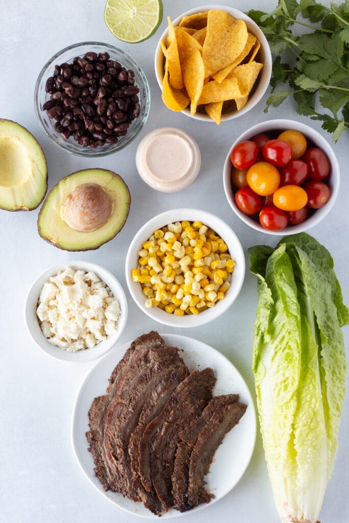 Top down shot of ingredients for a steak taco salad on a light gray surface. Ingredients include cooked steak, romaine lettuce, corn kernels, cherry tomatoes, avocado, black beans, cheese, and tortilla chips.