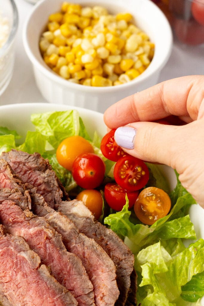 Close up of a hand placing cut cherry tomatoes in a bowl with steak and lettuce in it.