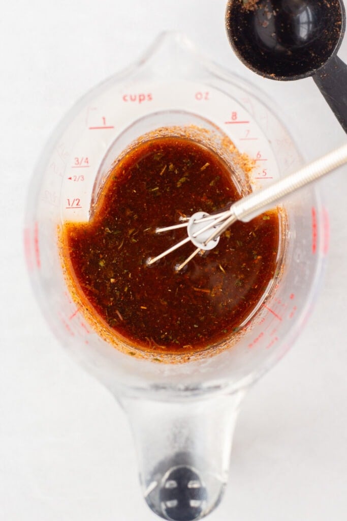 Top down close up of a measuring cup with a red taco marinade in it. A small whisk is in the cup as well, which rests on an off-white surface.