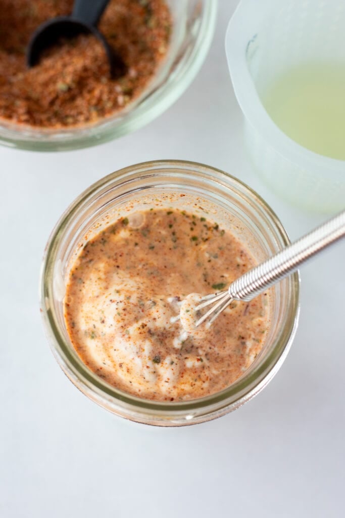 A small whisk stirring a dressing in a small mason jar made from lime juice, taco seasoning, and sour cream.