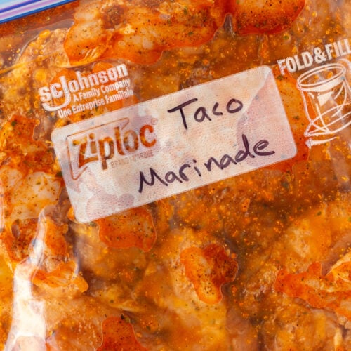 Top down shot of a plastic gallon sized ziploc bag holding chicken in a taco marinade. The words "taco marinade" are written in blank ink on top of the bag.