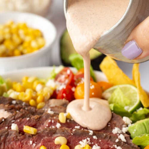 Close up of taco salad dressing being poured over a steak taco salad with bowls of ingredients shown in the background.