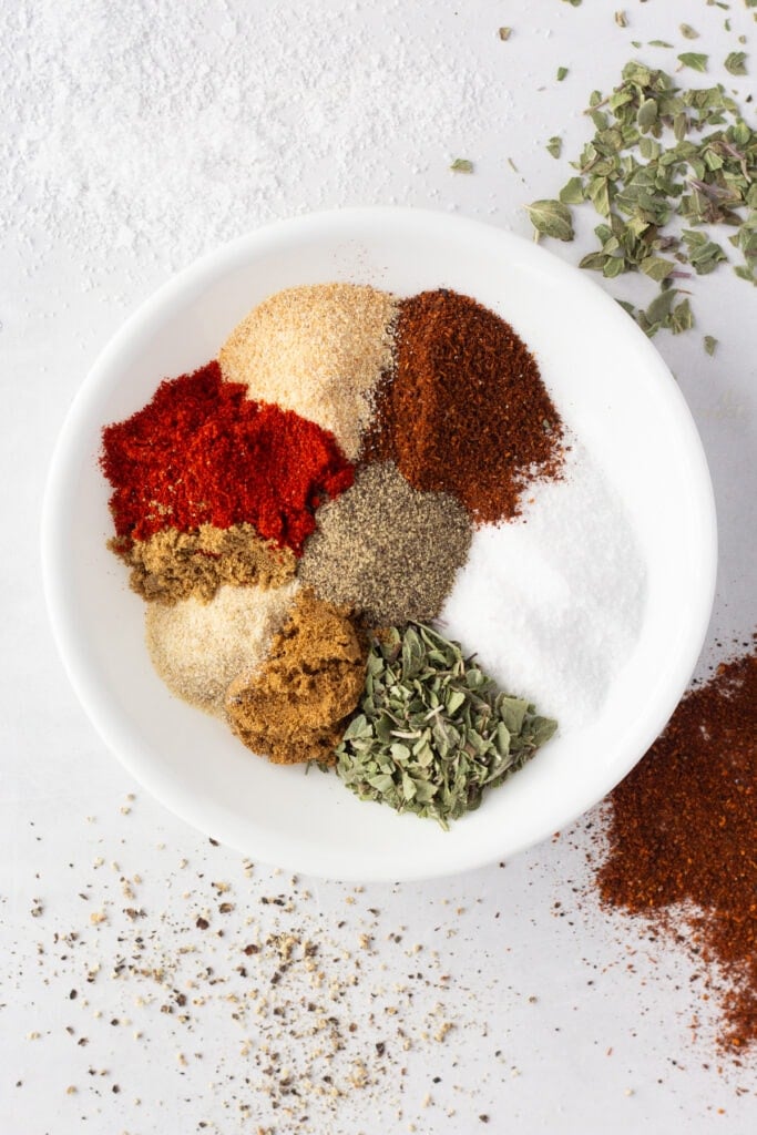Top down shot of 9 different spices bunched together on a small white dish. There are spices dashed on the white surface the dish is on as well.
