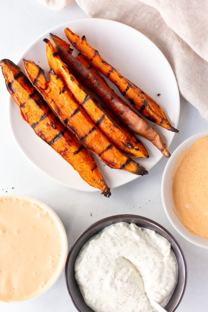 Top down shot of a small white plate with grilled sweet potato wedges on it next to three bowls with different dipping sauce in them. A tan cloth napkin is next to the plate and everything rests on a light gray background.