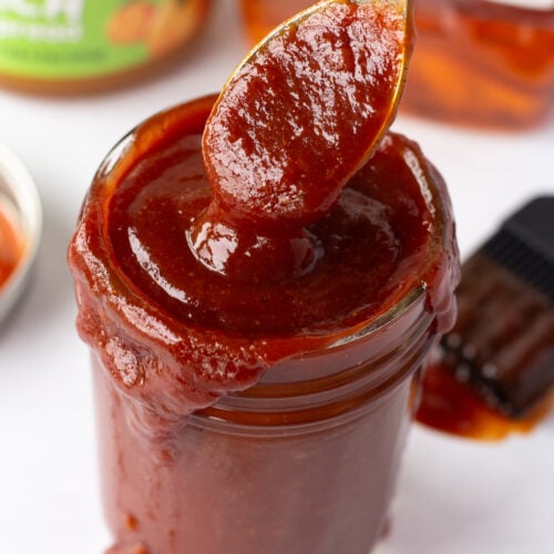 A jar with bourbon peach bbq sauce in it and a spoon dipping into it as well, with a lid, a bbq brush, a jar of peach preserves, and a bottle of jim beam in the background.