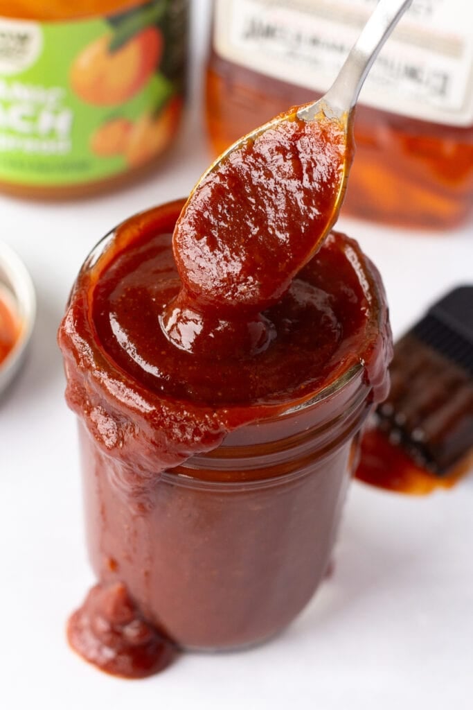 A jar with bourbon peach bbq sauce in it and a spoon dipping into it as well, with a lid, a bbq brush, a jar of peach preserves, and a bottle of jim beam in the background.