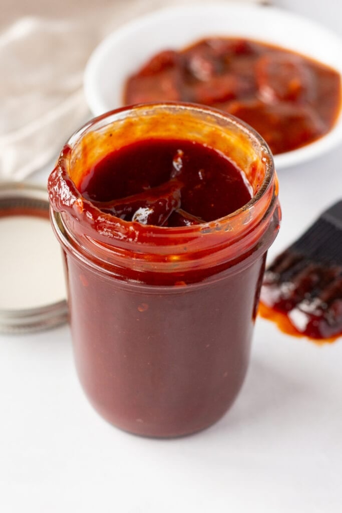 Chipotle bbq sauce in a small mason jar with a lid, a bbq brush, a plate of chipotle peppers, and a tan cloth napkin in the background.
