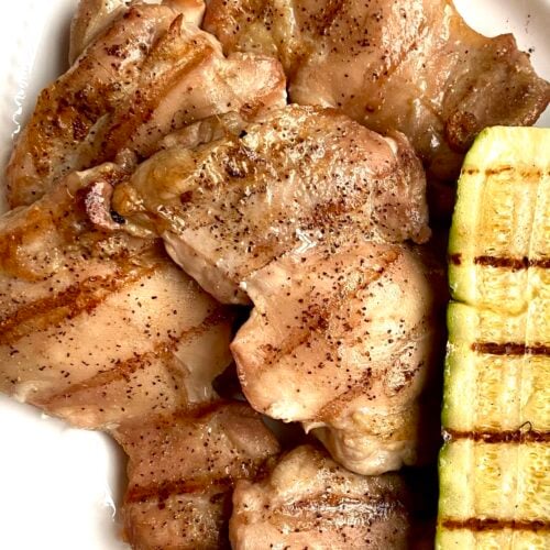 Close up of grilled boneless skinless chicken thighs on a white platter next to grilled zucchini.