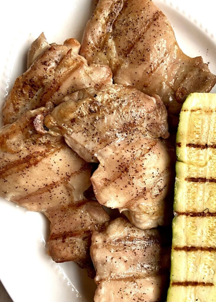 Close up of grilled boneless skinless chicken thighs on a white platter next to grilled zucchini.