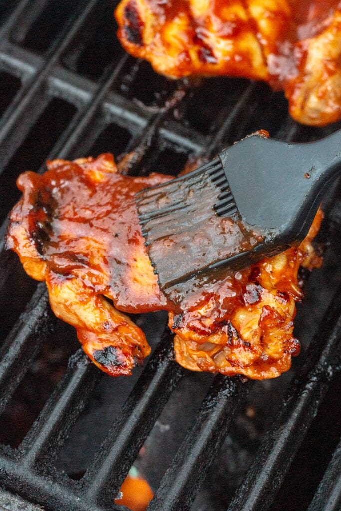BBQ boneless skinless chicken thighs on a grill being brushed with bbq sauce.