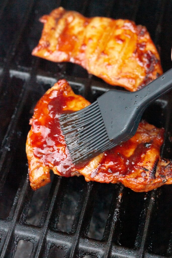 Boneless skinless chicken thighs on a grill being brushed with chipotle bbq sauce.