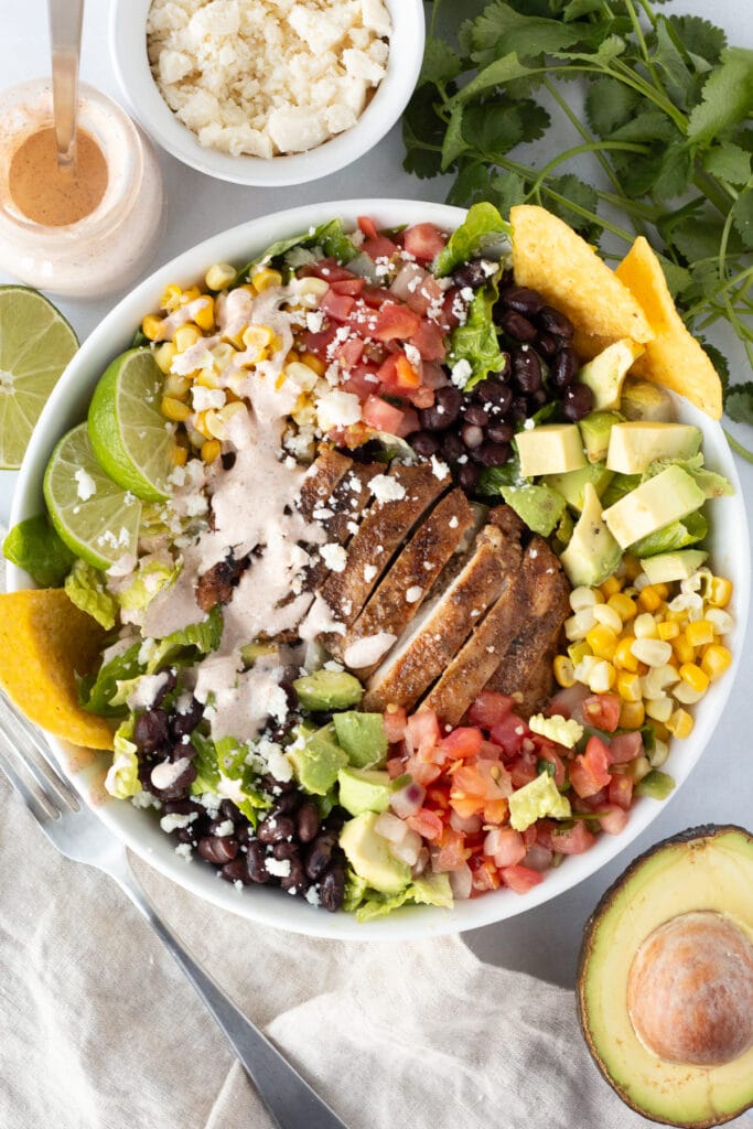 Top down shot of a grilled chicken taco salad in a white bowl with the ingredients surrounding it, like half an avocado, a small bottle of dressing, and some queso fresco cheese in a bowl.