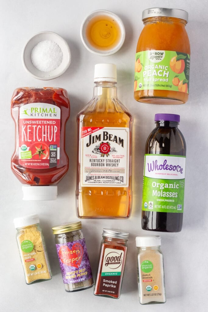 Top down shot of ingredients for peach bourbon bbq sauce on a light gray background, including ketchup, jim beam bourbon, molasses, peach preserves, vinegar, and spices.