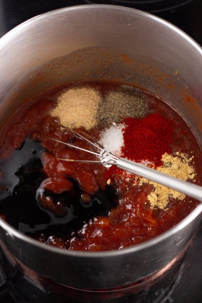 A sauce pan on a stove top with ketchup, molasses, spices, and jam in it, and a small whisk resting in it as well.