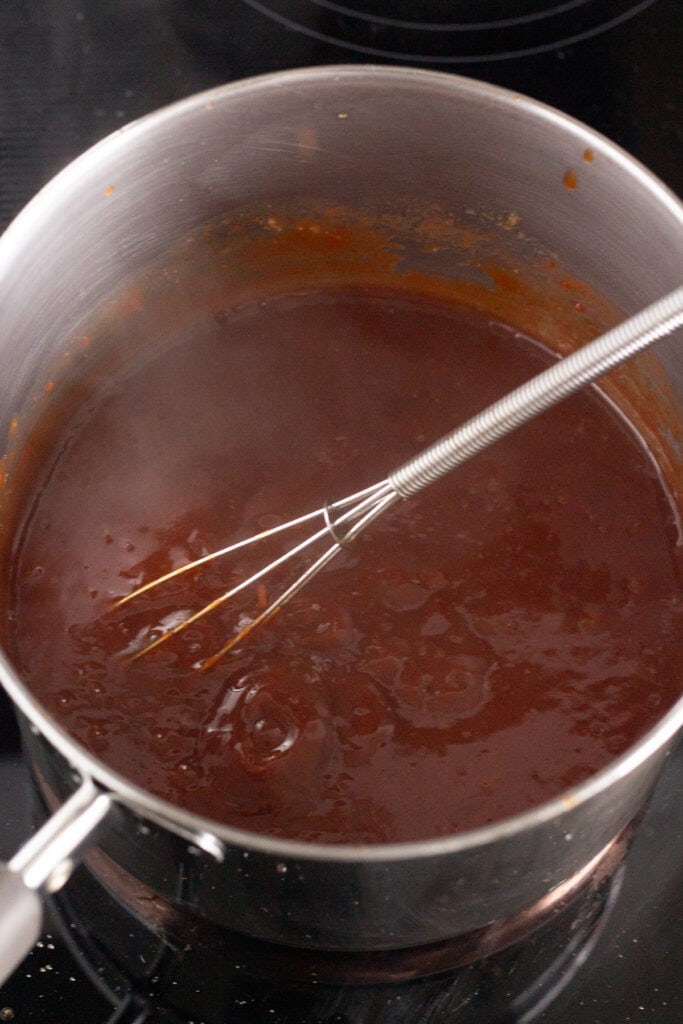 A sauce pan with BBQ sauce bubbling in it and a small whisk sticking out of it.