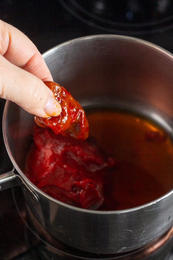 A hand putting a canned chipotle pepper into a saucepan on a stove top.