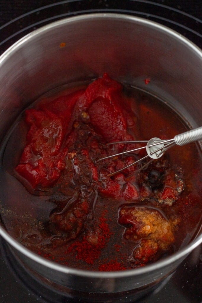 Close up of a small whisk stirring bbq sauce ingredients in a sauce pan, including tomato paste and maple syrup.