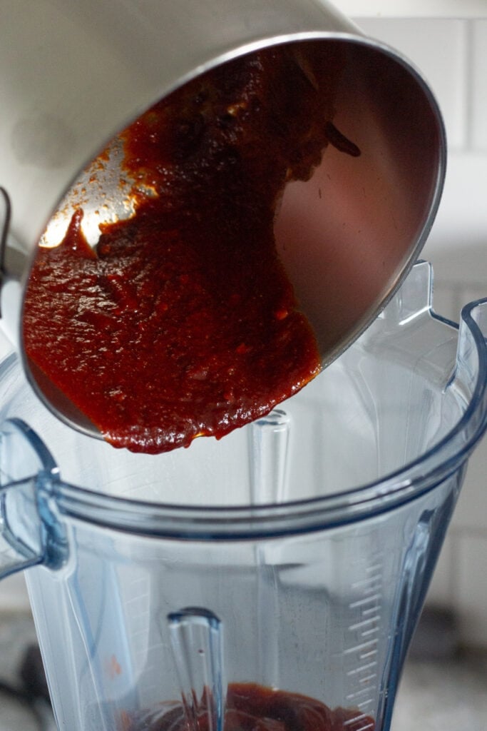 A red sauce being poured from a saucepan into a blender.
