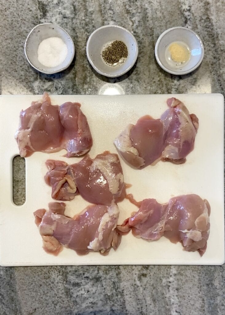 Top down shot of ingredients for grilled boneless skinless chicken thighs, including 5 chicken thighs on a white cutting board and small gray bowls with salt, pepper, and garlic powder in them.