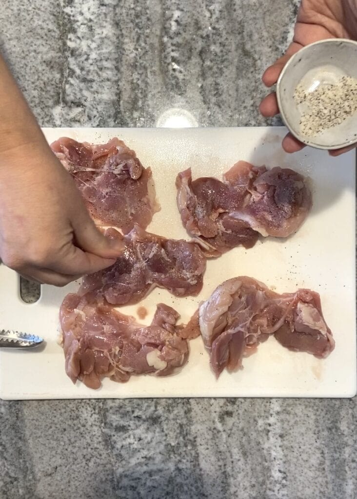 Top down shot of a hand seasoning raw boneless skinless chicken thighs on a white cutting board.