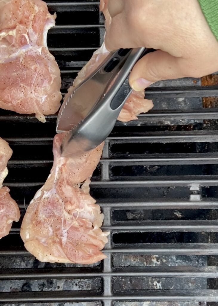 Close up of raw chicken  seasoned boneless skinless chicken thighs being placed on a hot grill with tongs.