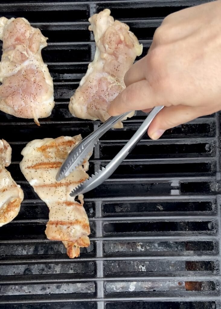 Close up of boneless skinless chicken thighs being flipped over on a hot grill.