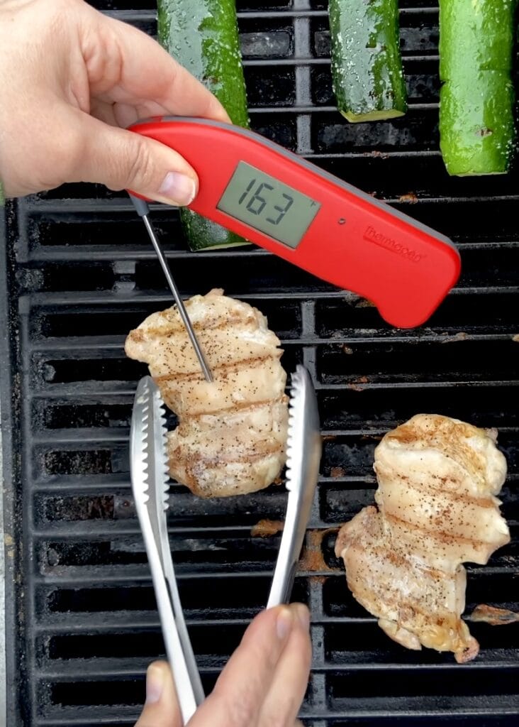 Boneless skinless chicken thighs being cooked on a hot grill next to zucchini, with a red meat thermometer sticking out of one thigh that reads 163F.