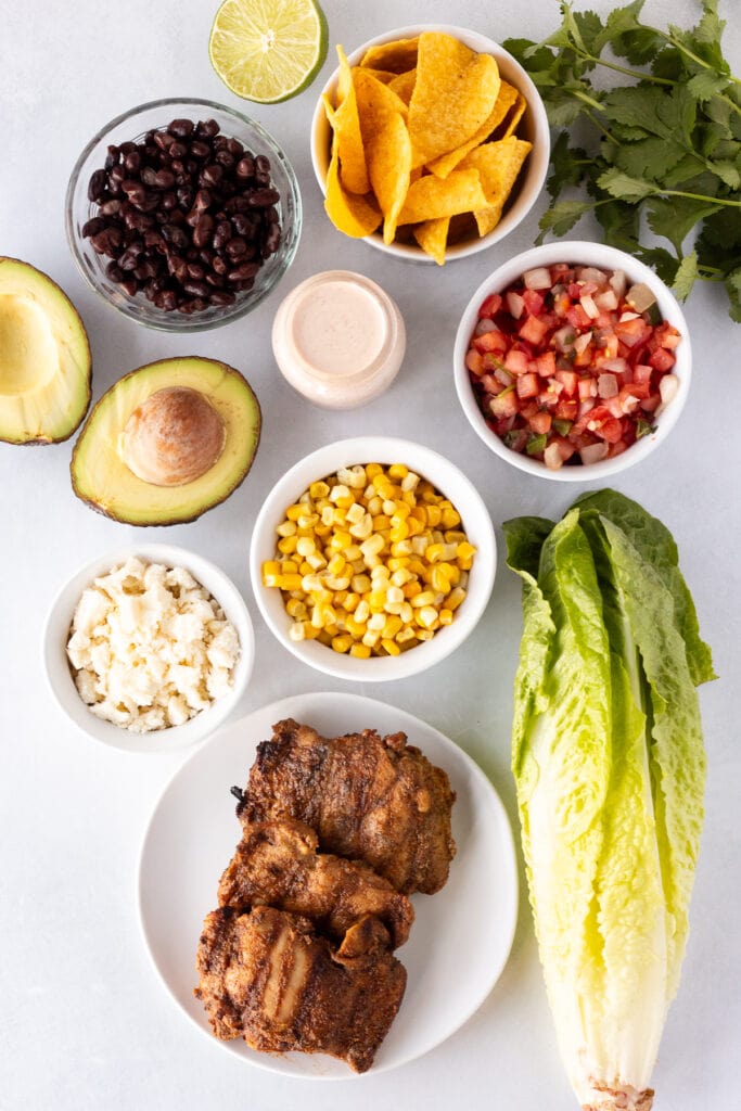 Top down shot of ingredients for a taco salad with grilled chicken on a white surface, including romaine lettuce, corn, pico de gallo, grilled chicken, avocado, black beans, cheese, and tortilla chips.