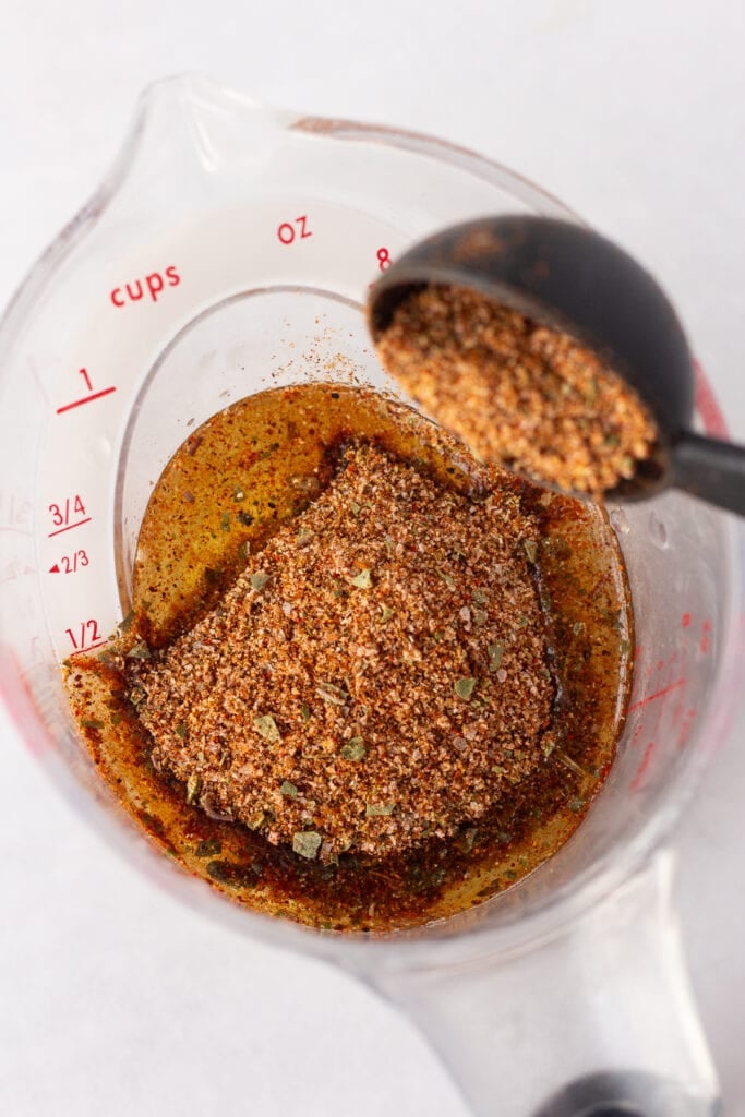 A taco seasoning blend being dumped into a plastic liquid measuring cup with oil and lime juice already in it.