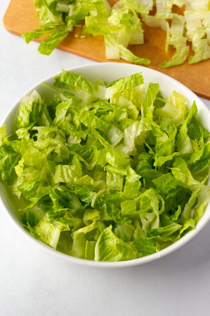 Chopped romaine lettuce in a white bowl with a wood cutting board and more romaine lettuce in the background.