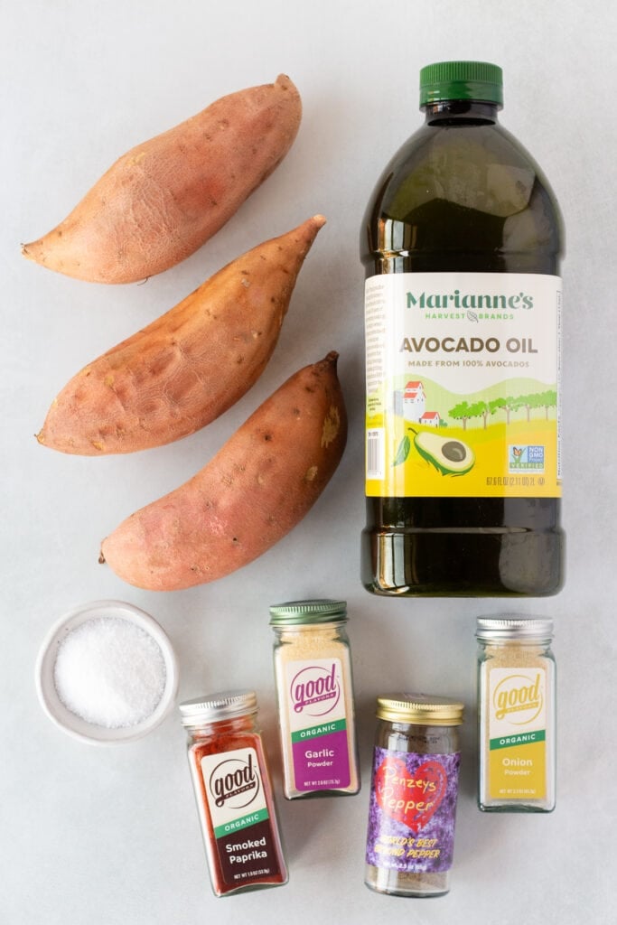 Top down shot of ingredients for grilled sweet potatoes, including a bottle of avocado oil, three sweet potatoes, and spice bottles.