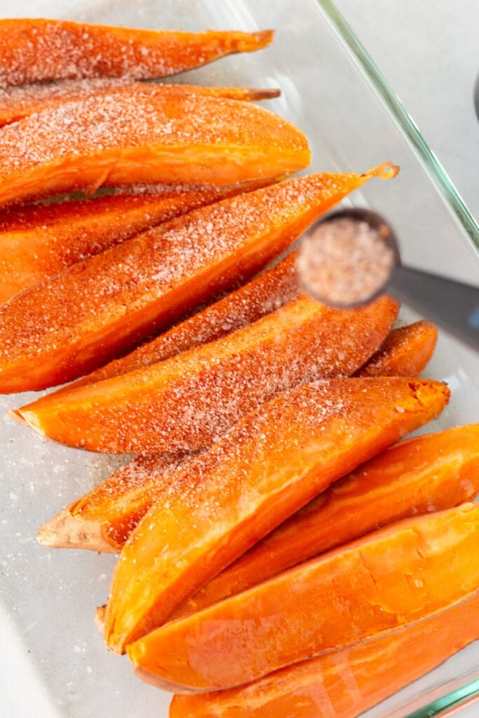 Uncooked sweet potato wedges in a rectangular clear dish being sprinkled with a spice seasoning.