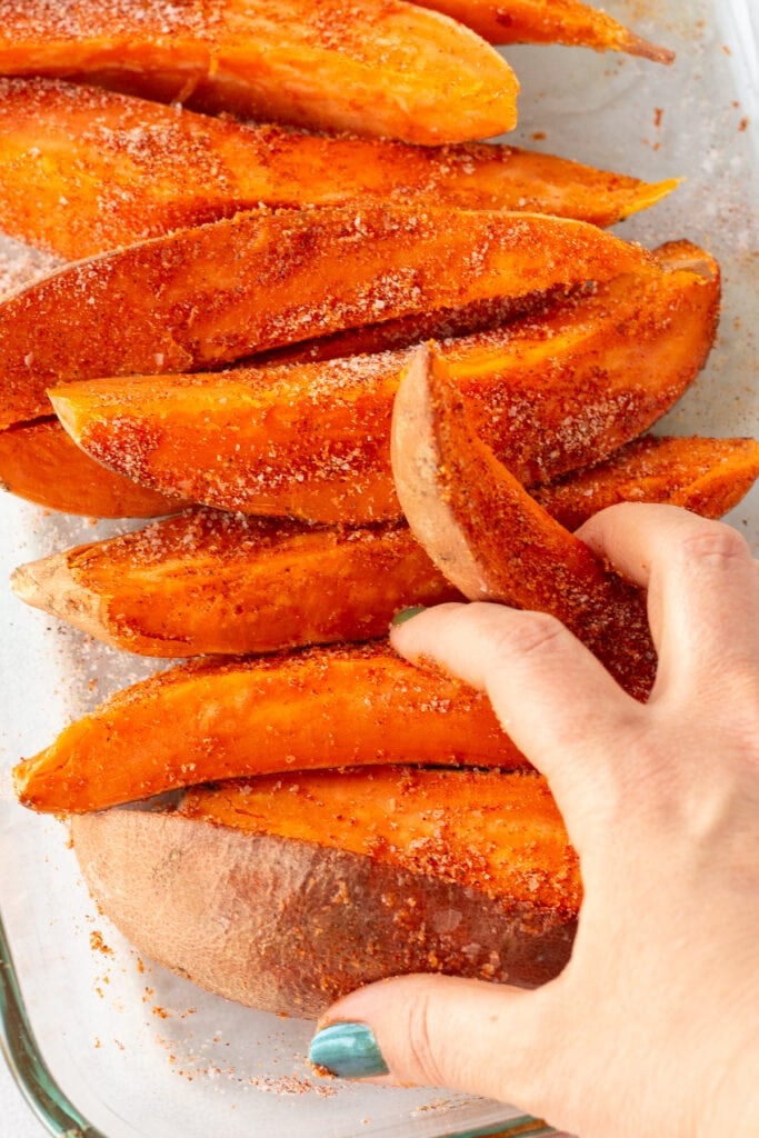Uncooked sweet potato wedges in a clear glass dish being stirred with by a pair of hands to coat in some oil and spices.