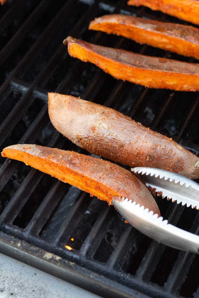 Close up of sweet potato wedges cooking on a grill grate and about to be turned over by a pair of metal tongs.