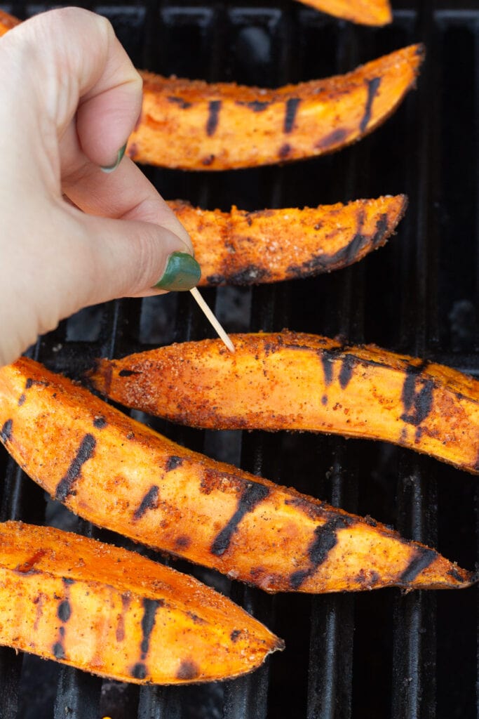 A hand pushing a toothpick into sweet potato wedges on a grill to test for doneness.