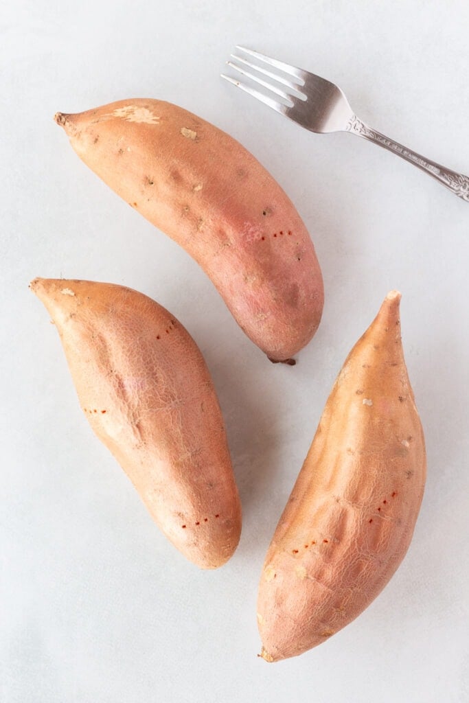 Three sweet potatoes with holes poked in them lying on a light gray background next to a fork.