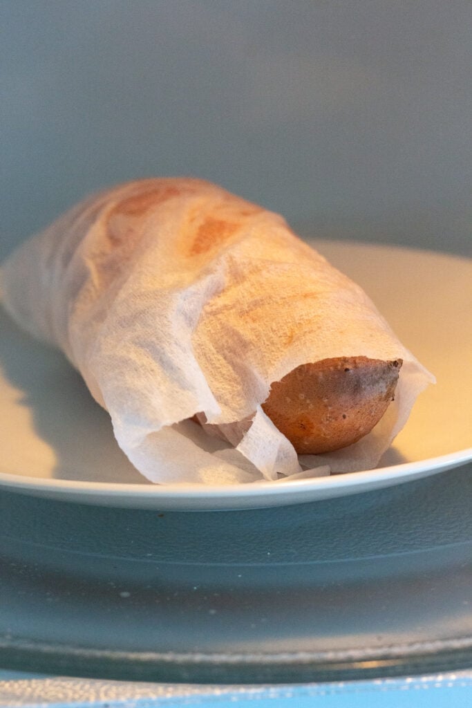 A sweet potato wrapped in a damp paper towel on a plate in the microwave.