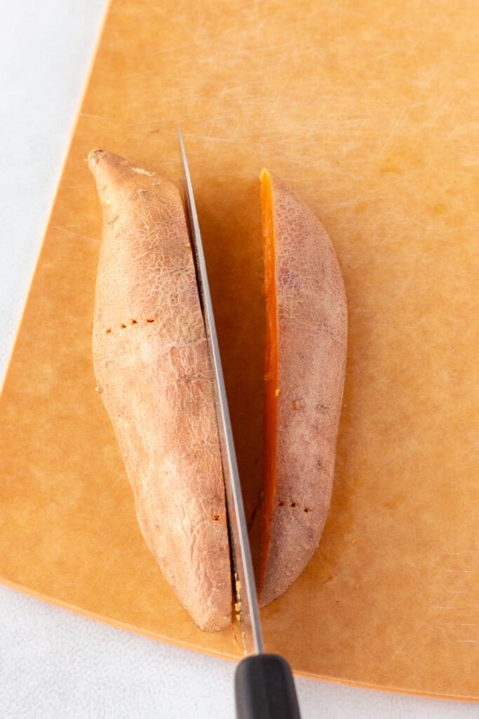 A large knife cutting half a sweet potato into half again on a wood cutting board.