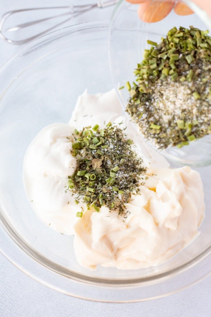 Pouring an herb and spice blend into a large clear bowl with mayo, sour cream, and Greek yogurt already in it.