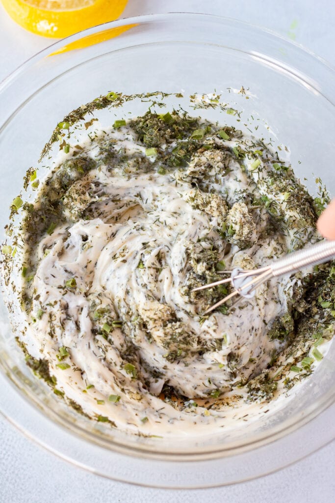 A small whisk stirring together homemade ranch dip in a large clear bowl.