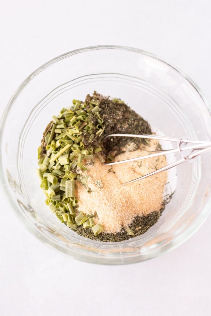 A small whisk stirring herbs and spices for a ranch seasoning together in a small glass bowl.