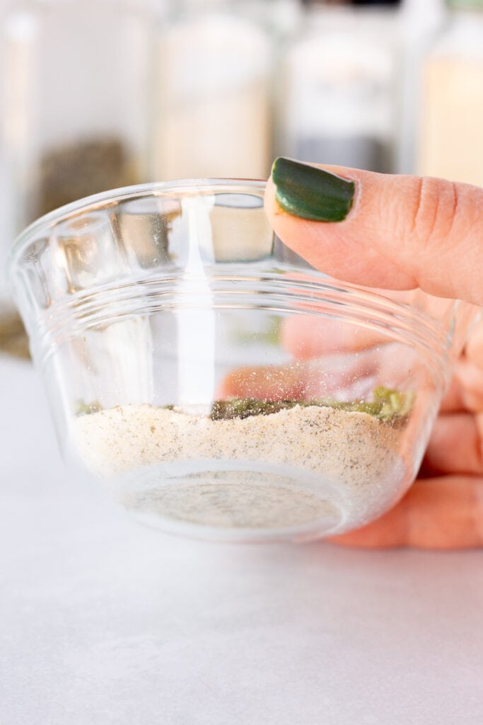 A small glass bowl with ranch seasoning mix in it being tipped up to show the bottom where the garlic and onion powder are resting.