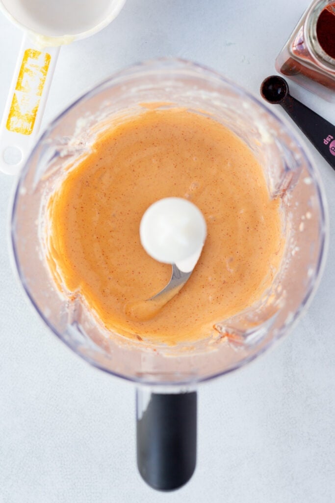 Top down shot of an orange smoked paprika mayo sauce in a blender on a light gray background.