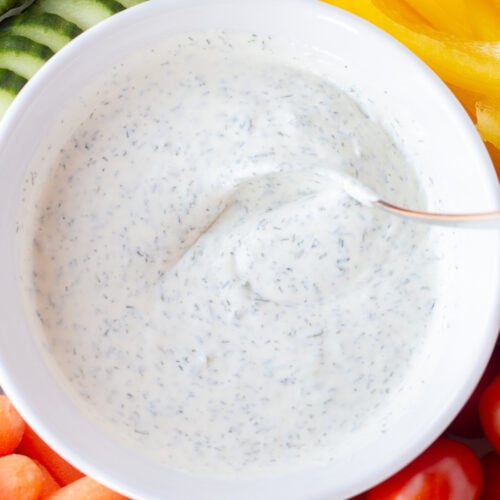 Top down close up of a small spoon swirling ranch dip in a white bowl that's embedded between cut cucumber, yellow bell pepper, cherry tomatoes, and baby carrots.