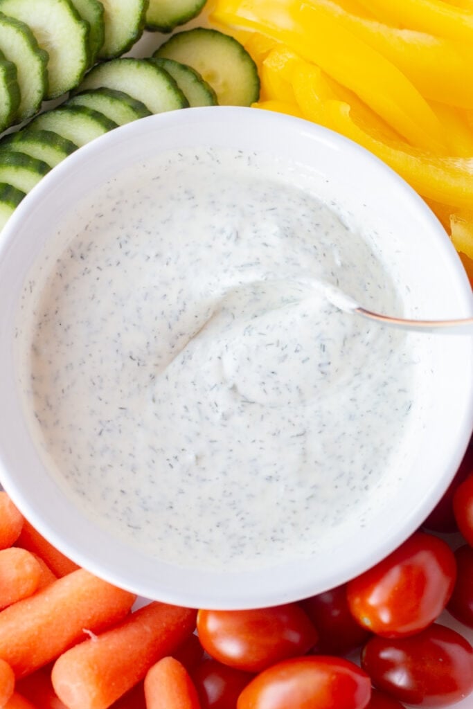 Top down close up of a small spoon swirling ranch dip in a white bowl that's embedded between cut cucumber, yellow bell pepper, cherry tomatoes, and baby carrots.