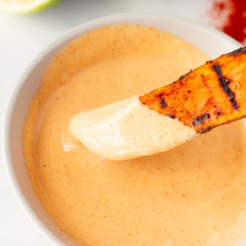 Close up of a grilled sweet potato wedge being dipped in a small bowl with an orange smokey mayo sauce in it.