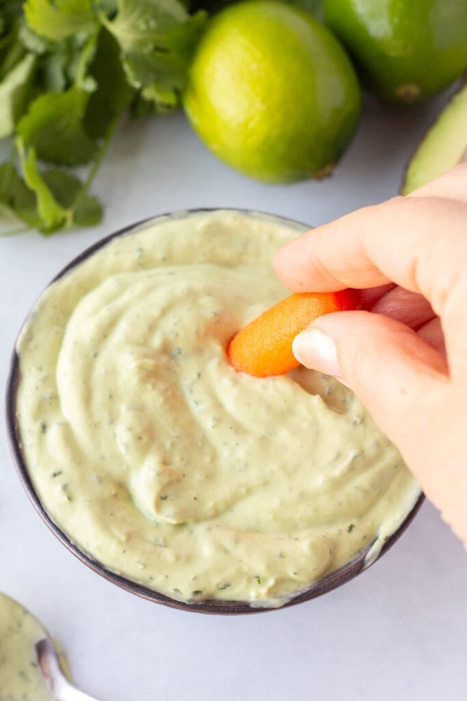 A hand dipping a baby carrot into a bowl with a green sauce in it. The bowl is surrounded by limes, fresh cilantro, half an avocado, and a spoon with the sauce on it.