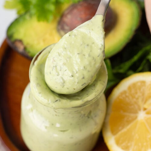 Close up of a jar of green avocado ranch dressing with a spoon scooping some out. The jar is on a brown wood plate with half an avocado, some fresh parsley, and half a lime on it as well.