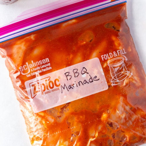 Top down shot of a ziploc bag with raw boneless chicken thighs in a red bbq marinade in it, with a small bowl of kosher salt and a jar of bbq sauce at the top of the frame.